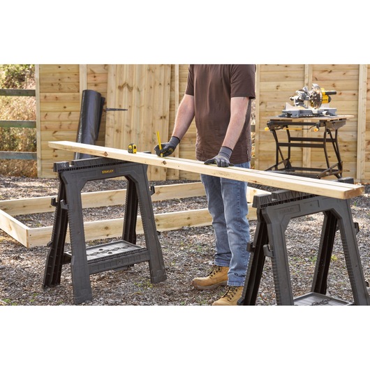 STANLEY 0-30-686 sawhorses supporting wood plank outdoors with person measuring, construction site setting.