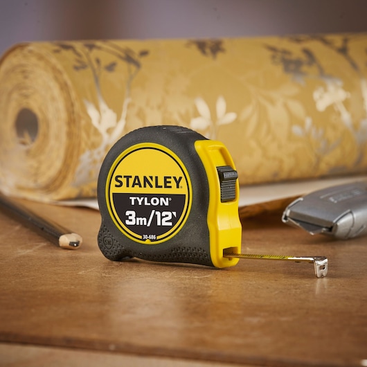 STANLEY 0-30-686 tape measure, 3m/12ft, black and yellow, shown on a work surface with wallpaper roll.