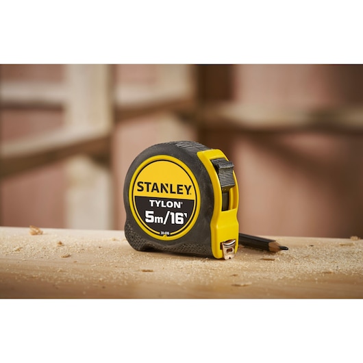 STANLEY 0-30-696 tape measure, yellow and black, 5m/16ft, side angle, on dusty wooden surface.