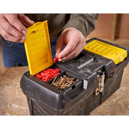 A person opening a yellow compartment on a black STANLEY toolbox, showing red wall plugs and metal screws organized inside.