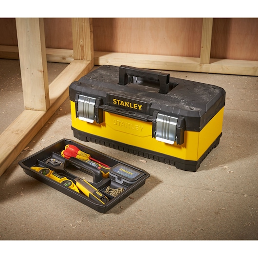 A yellow and black STANLEY toolbox with metal latches, placed on the floor near a wooden frame. The removable tray is positioned in front of the toolbox and contains various tools, including a hammer, screwdrivers, a spirit level, a tape measure, and screws.
