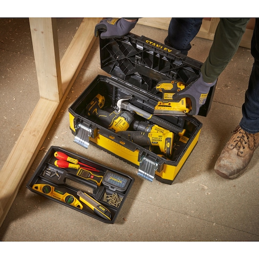 A person wearing gloves is opening a yellow and black STANLEY toolbox filled with various tools, including a drill, screwdriver, spirit level, and other hand tools, arranged neatly inside. The toolbox is placed on a workshop floor next to wooden beams.