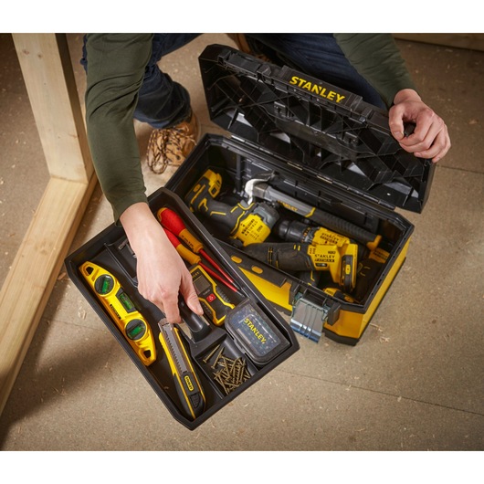 A person is opening a STANLEY tool box filled with various tools including a spirit level, utility knife, screwdriver, measuring tape, drill, and other hand tools. The box is organized with a removable tray containing smaller tools and screws.