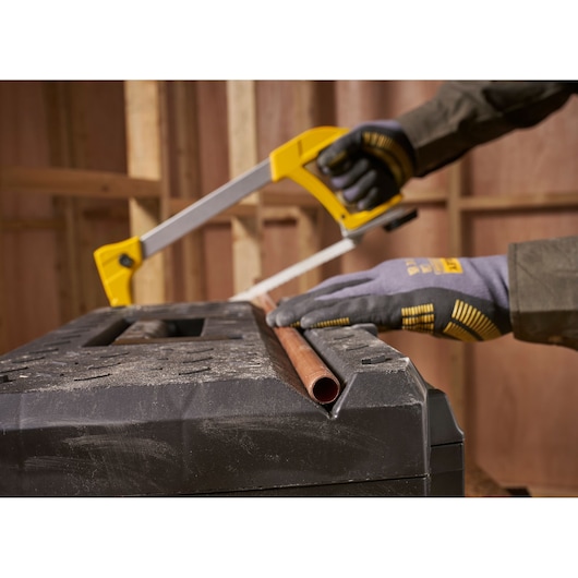 A person wearing gloves uses a yellow-handled hacksaw to cut a copper pipe, which is placed on a black, textured work surface in a construction setting.