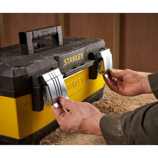 Hands opening the metal latches of a yellow and black STANLEY toolbox placed on a workbench with wood shavings.
