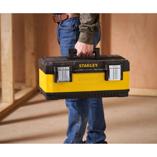 A person holding a STANLEY yellow and black toolbox with metal latches, standing in a wood-framed room.