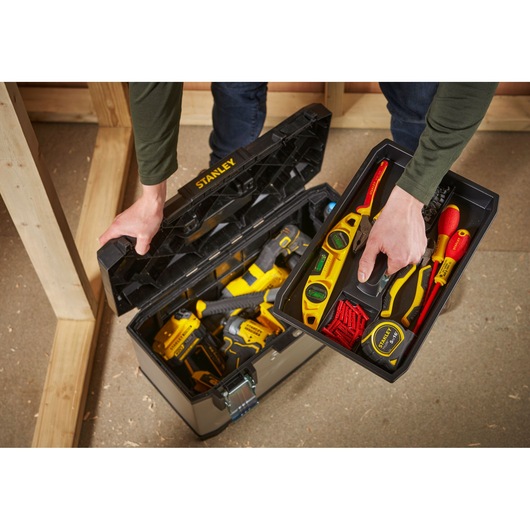 A person opens a STANLEY toolbox containing various hand tools and power tools. The removable tray holds items like a spirit level, screwdrivers, a utility knife, and a box of screws. The main compartment contains larger tools including a drill.