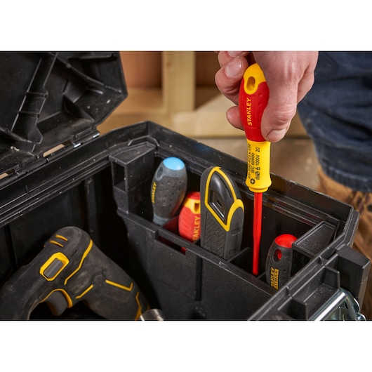 A close-up of a person's hand holding a STANLEY insulated screwdriver above an open black toolbox that contains various tools, including screwdrivers and a drill.