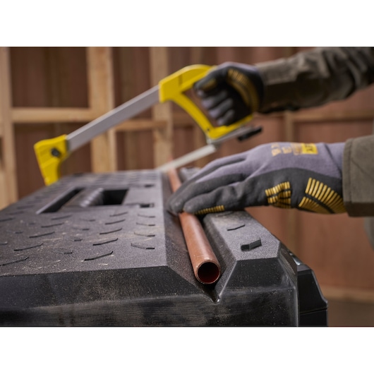 A person wearing work gloves uses a yellow hacksaw to cut a copper pipe on top of a black textured toolbox.