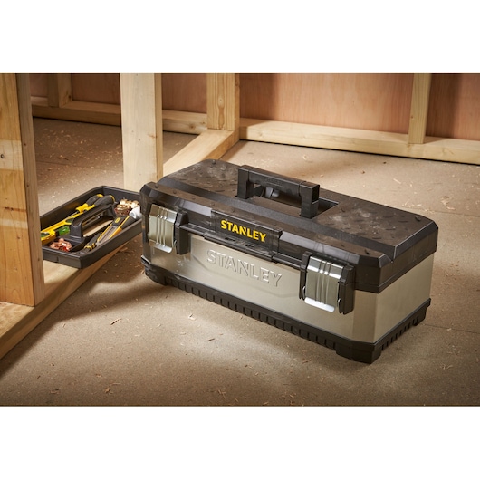 A STANLEY metal and plastic toolbox with handle and secure latches is placed on a workshop floor. The toolbox tray with various hand tools is visible beside it.