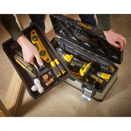 A STANLEY toolbox with various yellow and black hand tools, including screwdrivers, a spirit level, utility knife, wrench, and plumbing fittings. Some power tools are visible inside the open toolbox. The scene shows a person lifting the tray out of the toolbox.