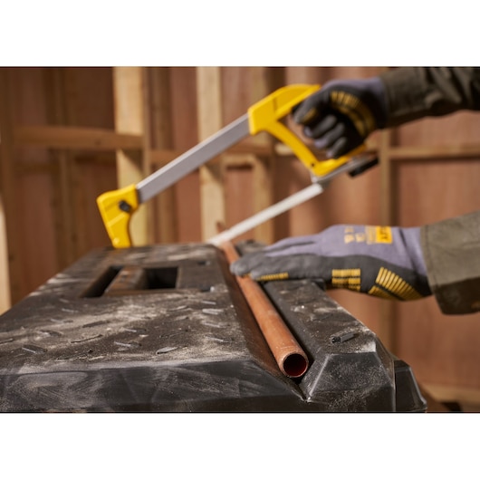 A person wearing STANLEY branded gloves uses a yellow-handled hacksaw to cut a copper pipe on a workbench in a construction setting.