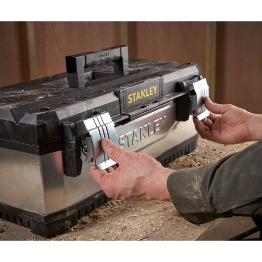 Hands opening the metal latches of a STANLEY toolbox placed on a workbench, with wood shavings around.