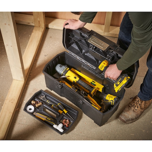 A person opens a black STANLEY plastic tool box filled with various hand tools and power tools. The removable tray holds small tools and accessories, while the main compartment contains larger tools. The tool box is being used on a construction site with wooden framing.