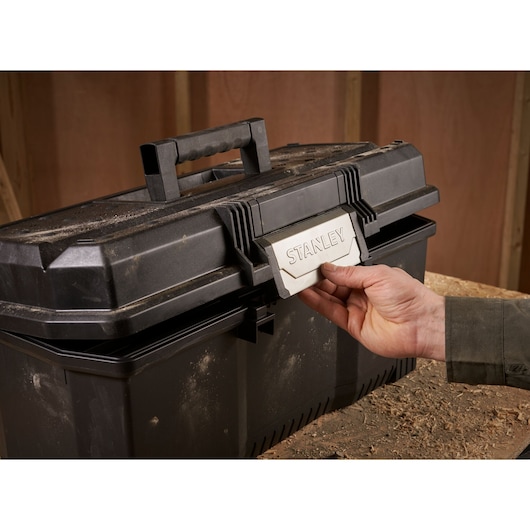 A hand opening a black STANLEY toolbox with dust and wood shavings on the surface, placed on a workbench.