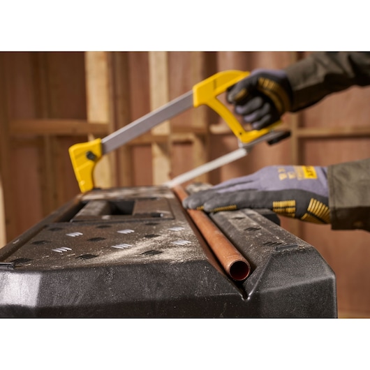 A person wearing gloves uses a yellow-handled hacksaw to cut a metal pipe placed on a black workbench in a workshop setting.