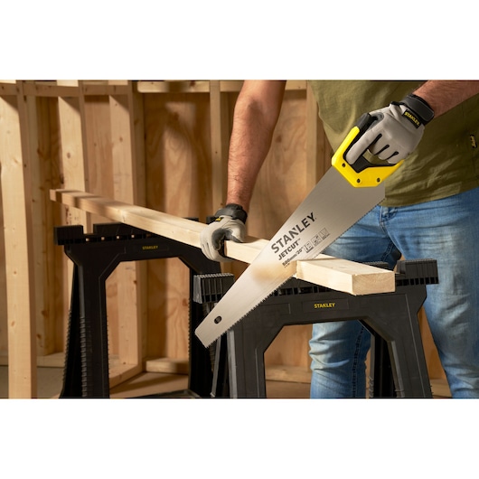 STANLEY 2-15-288 hand saw with yellow handle cutting wood plank on black sawhorse, workshop setting.