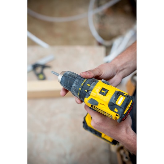 Photo of STANLEY FMC607B cordless drill driver held in hand, yellow and black design, workshop background.