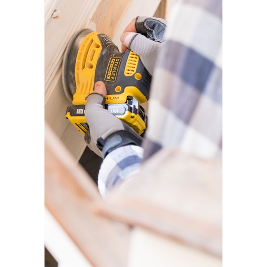 STANLEY FMCW220B cordless sander in use on wood, close-up showing yellow tool and gloved hands.