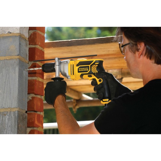 STANLEY FME140K hammer drill in use, drilling into brick wall, yellow and black body, user wearing gloves.