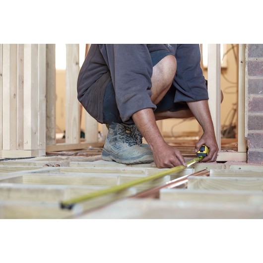 STANLEY FMHT0-36327 tape measure in use by a worker measuring wooden floor framing at a construction site.