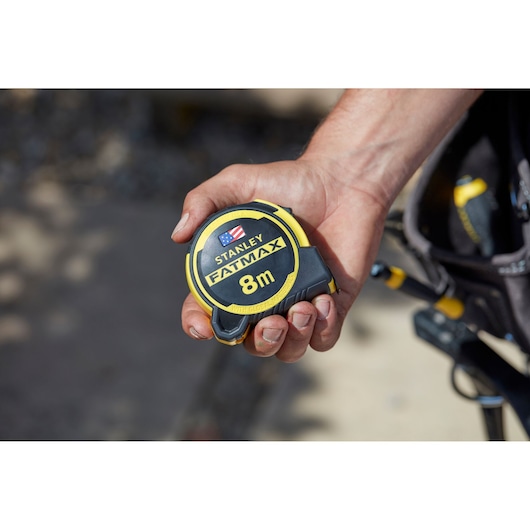 Photo of a STANLEY FATMAX 8m tape measure, SKU FMHT0-36327, held in a person's hand on a job site.