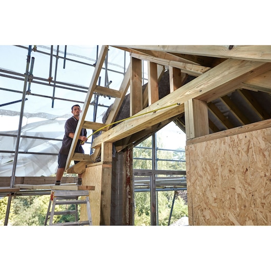 STANLEY FMHT0-36327 tape measure in use at timber construction site, shown in action measuring wooden beams.