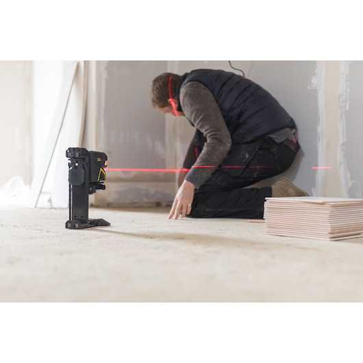 STANLEY FMHT1-77415 laser level projecting red line in construction site with worker and plywood sheets.
