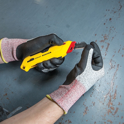 Stanley FMHT10362 utility knife held by gloved hands, close-up angle, yellow body with black details.