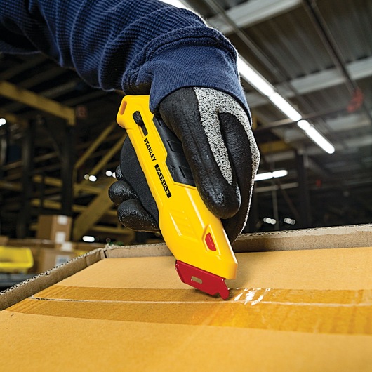 Stanley FMHT10362 yellow utility knife cutting box tape, work glove hand, warehouse background.