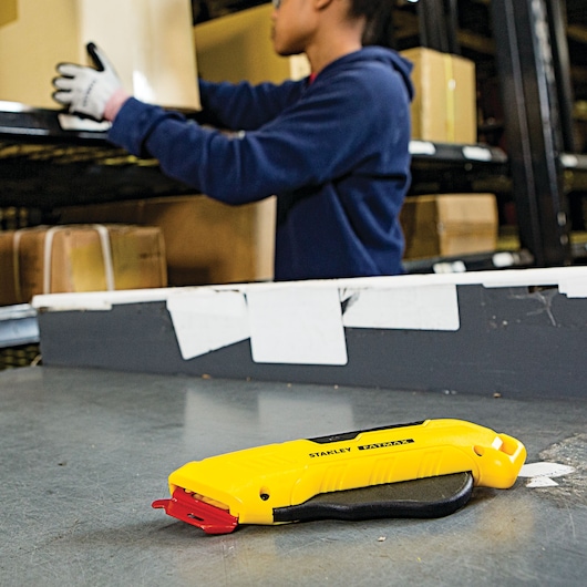 STANLEY FMHT10362 yellow safety utility knife on warehouse table, person handling boxes in background.