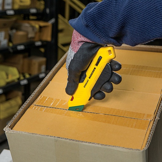 Stanley FMHT10363 yellow safety utility knife cutting box tape, close-up handheld in warehouse setting.