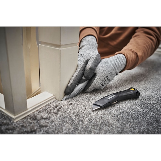 Photo of STANLEY FMHT10501-0 utility knife being used to cut carpet along base of white post, with gloves.