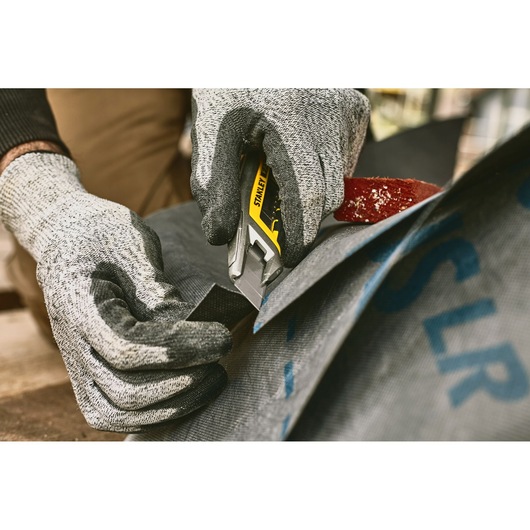 Photo of STANLEY FMHT10594-0 utility knife cutting fabric, close-up with gloved hands.