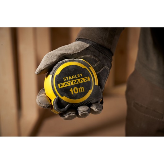 STANLEY FMHT33005 10m FATMAX tape measure held in a gloved hand, close-up indoor setting.