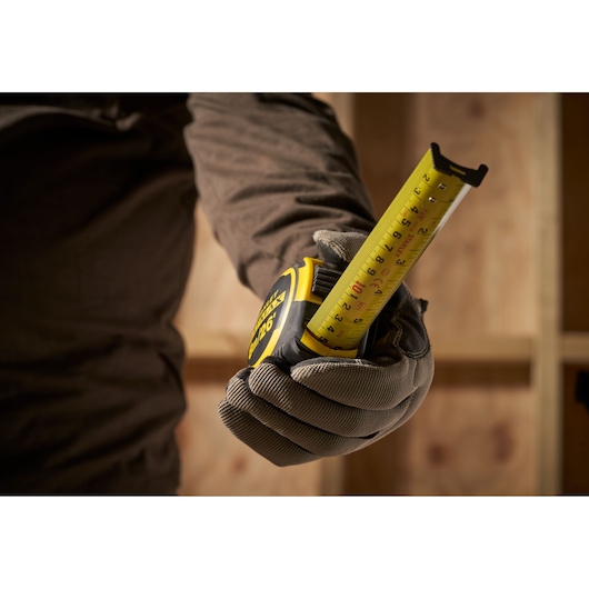 Photo of STANLEY FMHT33105 tape measure held by gloved hand, yellow blade extended, woodworking background.