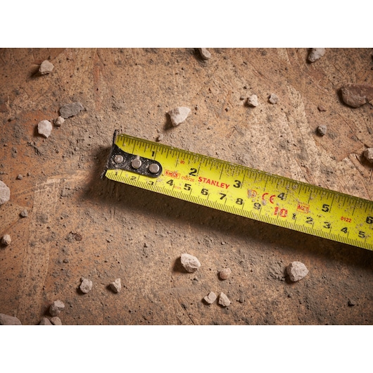 STANLEY FMHT33105 yellow tape measure extended on a dusty work surface with small rocks, top view.