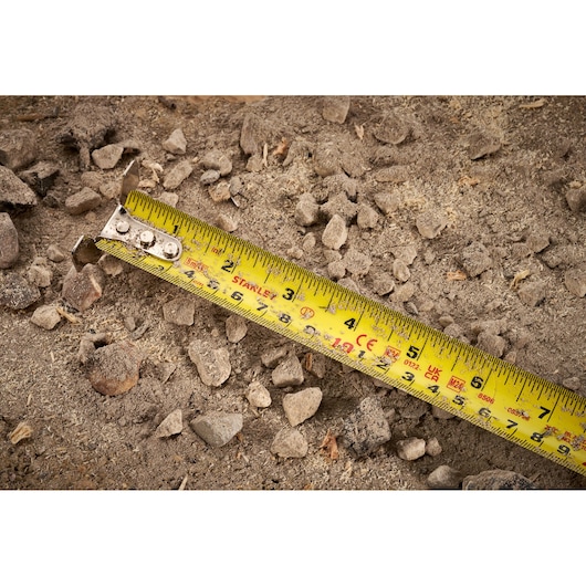 STANLEY FMHT38215-5 yellow tape measure on dirt and rocks, close-up photo.