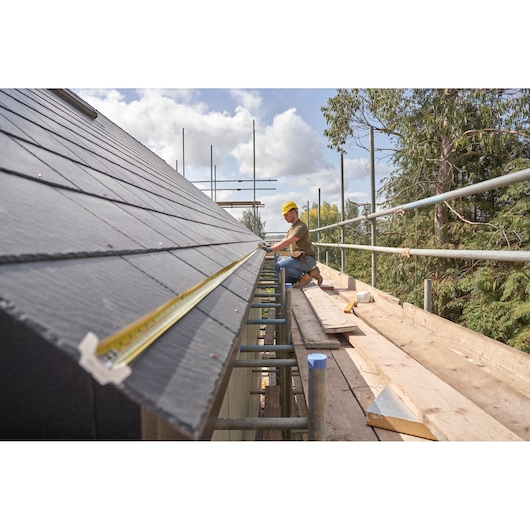 STANLEY FMHT38218-5 tape measure extended along a roof edge with worker on scaffolding, outdoor construction site.
