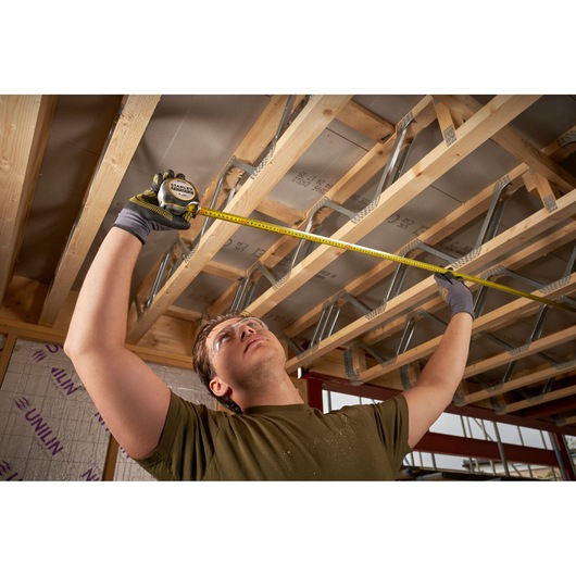 STANLEY FMHT38218-5 tape measure used by person wearing gloves to measure wooden ceiling beams in construction site.