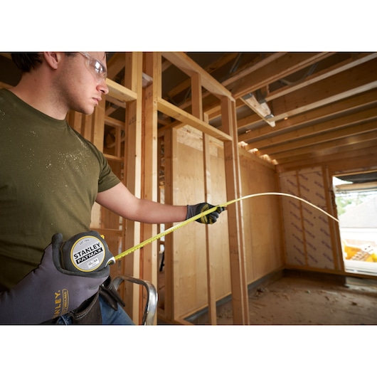 STANLEY FMHT38233-5 tape measure extended by worker in gloves at a wooden construction site.