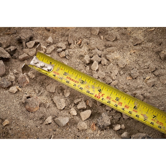 STANLEY FMHT38233-5 tape measure on rocky dirt surface, close-up showing yellow blade and markings.