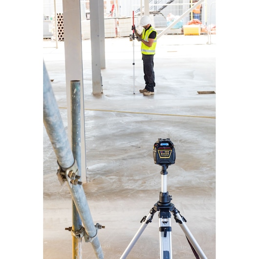 STANLEY FMHT77447-1 rotary laser level on tripod at a construction site, worker in safety gear in background.
