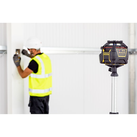 STANLEY FMHT77447-1 rotary laser level on tripod, construction setting, worker in background.