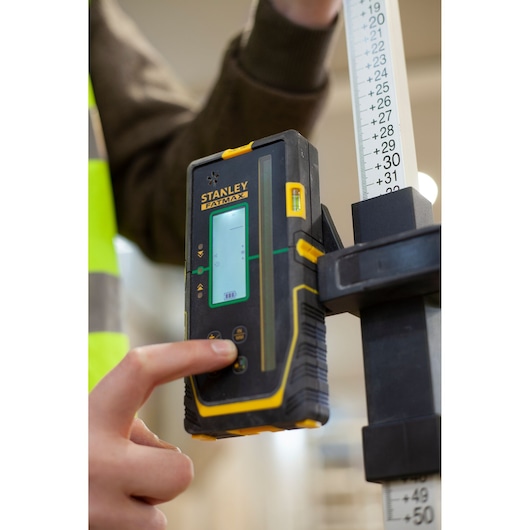 STANLEY FMHT77653-0 laser detector held by a person pressing a button, next to a measuring rod.