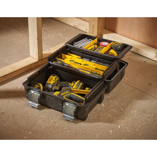 A black Stanley tool box with two compartments, open and filled with various yellow and black tools and accessories, including a drill, tape measure, screwdrivers, levels, and other hardware, placed on a workshop floor near wooden framing.