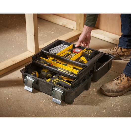 A person is opening a black Stanley tool box with metal latches, containing various yellow and black hand tools and accessories such as a level, tape measure, screwdriver, and drill bits. The tool box is being used on a workshop floor, surrounded by wooden construction.