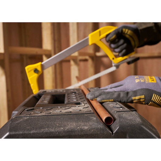 A person wearing protective gloves uses a yellow and silver STANLEY hacksaw to cut a copper pipe on a sturdy black workbench in a workshop setting.