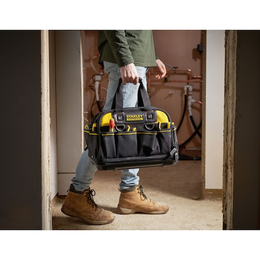 STANLEY FMST1-73607 tool bag with yellow accents, carried by person wearing work boots indoors.