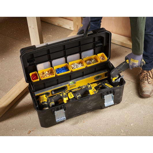 A STANLEY tool box with an open lid showing assorted tools, a level, and yellow containers filled with screws and connectors. A person wearing gloves is holding a STANLEY power tool, and the box is placed on a workshop floor.
